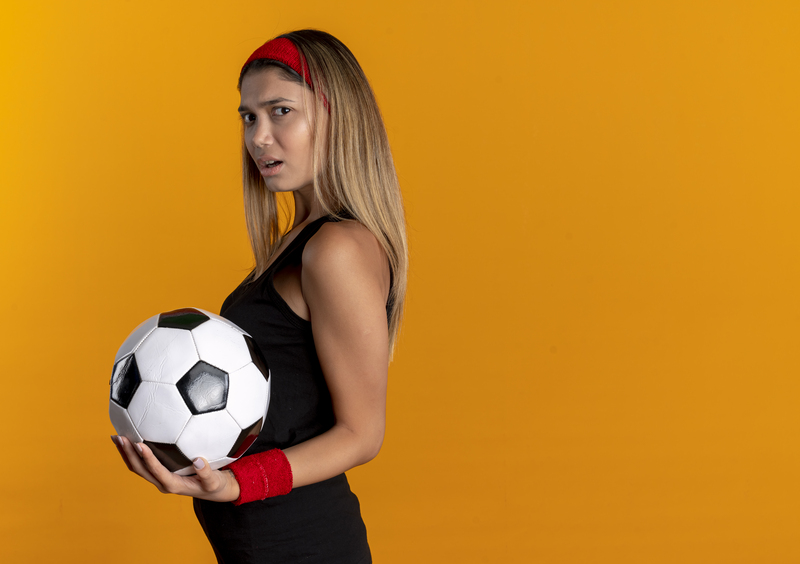 young-fitness-girl-black-sportswear-red-headband-holding-soccer-ball-displeased-standing-orange-wall.jpg